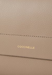 Beiges Leder mit strukturierter Oberfläche, ausgestattet mit einem goldgeprägten "COCCINELLE"-Logo und kontrastierenden Stichdetails.