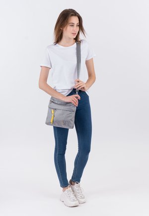 Woman in white t-shirt, blue jeans, and white sneakers stands holding gray crossbody bag with yellow tassel on front pocket.