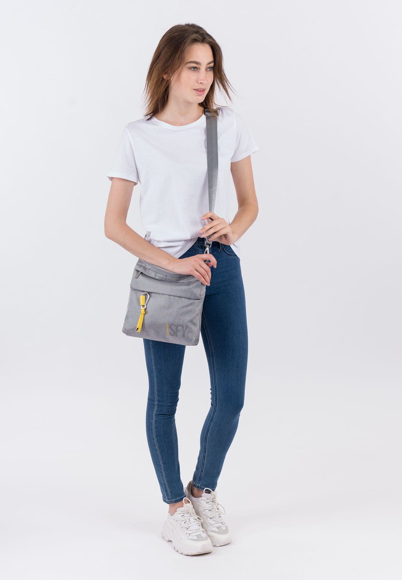 Kvinde i hvid T-shirt, blå jeans og hvide sneakers står og holder en grå crossbody taske med gul kvast på frontlommen.