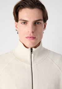 Vit zip-up jacka med hög krage, med ribbade detaljer och en mjuk textur. Närbild som fokuserar på framsidan av designen.