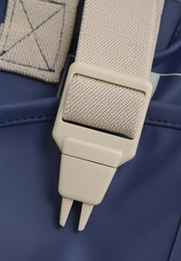 Marineblaue Trage mit einem strukturierten beigen Stoff und einer Kunststoffschnalle. Die Schnalle verfügt über Zacken zur Befestigung, was das funktionale Design verbessert.