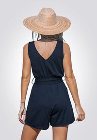 Jardín azul marino hecho de una tela suave, con un diseño de espalda en V y una cintura elástica. Combinado con un sombrero de paja de ala ancha.