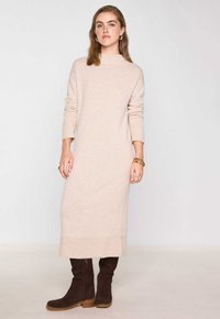Abito beige lavorato a maglia con scollatura alta, maniche lunghe e polsini a costine, abbinato a stivali al ginocchio in camoscio marrone e semplici accessori in oro.