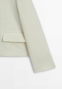 Blazer de couleur crème avec une texture douce et un design élégant. Il dispose d'une poche unique et d'une coupe ajustée. Fabriqué en tissu tissé doux.