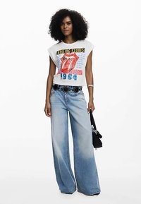 White, sleeveless cropped t-shirt featuring a Rolling Stones graphic, paired with light blue wide-leg jeans and a black belt with circular accents.
