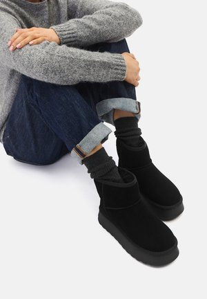 Schwarze Wildleder-Stiefeletten mit flacher Sohle, weicher Textur und seitlichem Ausschnitt. Getragen mit hochgekrempelter Jeans und dunklen Stricksocken.