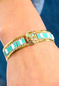 Bracelet doré avec des perles en turquoise et blanches, comportant un élément décoratif central. Texture lisse avec des détails intriqués.