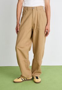 Beige wide-leg trousers made of cotton with side pockets, featuring a button closure, and a relaxed fit. Paired with yellow sporty shoes.