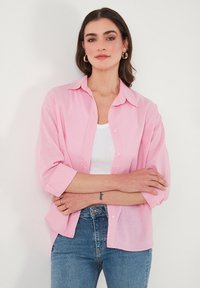 Light pink, button-up shirt with a collar, long sleeves rolled to elbows, paired with a white tank top and blue jeans. Smooth, lightweight fabric.