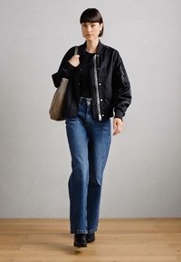 Chaqueta bomber negra con cremallera plateada, parte superior negra, jeans azules de cintura alta y botas negras hasta el tobillo; llevando un bolso tote gris claro.