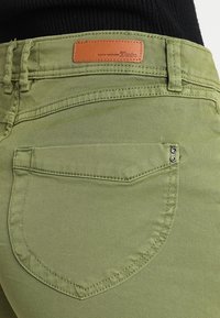 Jeans de color verde oliva con un diseño ajustado, detalles de costura, un pequeño bolsillo trasero y una etiqueta de cuero marrón con el nombre de la marca.