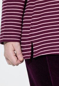 Top morado de manga larga con rayas horizontales rosas, que presenta aberturas laterales y un corte relajado, combinado con pantalones de pana morados y suaves.