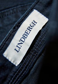 Valmis ”LINDBERGH” sinisellä tekstillä valkoisessa kangaslapussa, ommeltu tumman denimsiniseen materiaaliin näkyvin saumaratkaisuin.