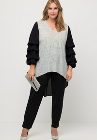 Mujer vestida con pantalones negros y una blusa blanca de manga larga con un patrón, mangas abullonadas negras, sosteniendo un clutch plateado y usando tacones plateados.