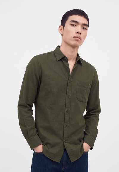 Jeune homme aux cheveux courts noirs portant une chemise olive à boutons avec manches longues et un pantalon bleu foncé, debout les mains dans les poches.