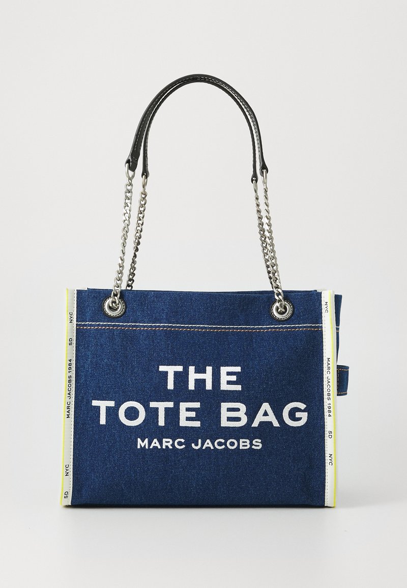 Mörkblå denim tote-väska med vit text "THE TOTE BAG MARC JACOBS", silverkedja och svarta läderhandtag samt vita sidokantdetaljer.
