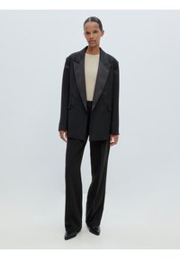 Zwarte dubbelbreasted blazer met satijnen revers, beige fitted top en zwarte broeken. Slank ontwerp, een zachte textuur en puntige hakken.