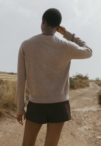 Beige stickad tröja med rund halsringning och långa ärmar, tillsammans med mörka shorts. Texturen verkar mjuk och lätt. Naturlig utomhusmiljö.