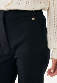 Czarne dopasowane spodnie o gładkiej tkaninie, z małym złotym logo na pasku, w połączeniu z kremowym swetrem o teksturowanym splotcie.