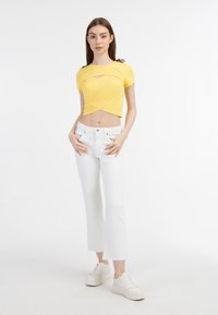 Sárga bordás crop top, keresztbe álló dizájnnal, fehér magas derekú farmernadrággal és fehér sportcipőkkel párosítva. Tiszta, minimalista stílus.