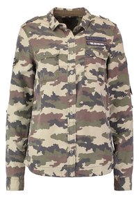 Chemise à manches longues boutonnée avec un motif camouflage vert, marron et beige et deux poches poitrine, dont l’une avec une étiquette visible « Superdry ».
