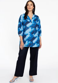 Blau gemusterte Bluse mit drei Viertelärmeln und V-Ausschnitt, kombiniert mit dunklen weiten Hosen. Blaue Riemchensandalen runden den Look ab.