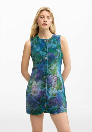 Szőke nő ujjatlan, gombos, kék-zöld batikolt mintás, virágmintás miniruhában, fehér, sima háttér előtt áll.