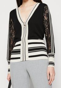 Kvinna iklädd svart- och vitrandig cardigan med spetsärmar, pärlhalsband och ljusgrå byxor som håller en svart handväska.