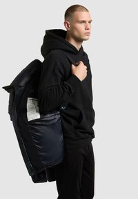 Zwarte hoodie en broek gedragen door een mannelijke figuur. Hij houdt een marineblauwe pufferjack aan met een glanzende textuur en een zichtbaar binnenlabel, tegen een grijze achtergrond.