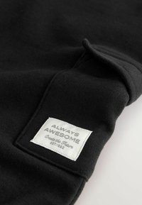 Schwarzer Sweatshirt-Stoff mit glatter Textur, der ein weißes Etikett zeigt, auf dem "ALWAYS AWESOME" und "Create the Future" steht.