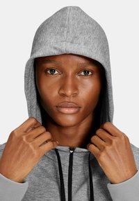 Grauer Kapuzenpullover mit Kordelzug, strukturierter Oberfläche, schwarzen Akzenten neben dem Reißverschluss und Rippbündchen. Das Modell hält die Kapuze mit beiden Händen.