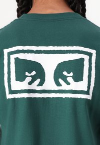 Πράσινο μπλουζάκι με κοντά μανίκια και λαιμόκοψη crew, διακοσμημένο με μια μεγάλη λευκή γραφική παράσταση χεριών μέσα σε ορθογώνιο πλαίσιο, από βαμβακερό υλικό.