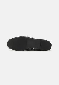 Baskets en maille noire avec une semelle en caoutchouc, présentant un motif texturé et un logo embossé sur le côté. Le dessous est plat avec des rainures pour une meilleure adhérence.