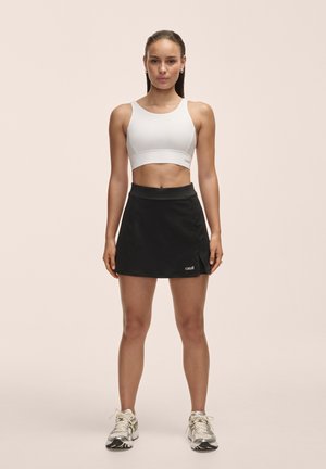Biały crop top i czarna sportowa spódnica, wykonane z gładkiego materiału. Spódnica ma logo i detal w postaci rozcięcia. Buty są srebrno-czarne.