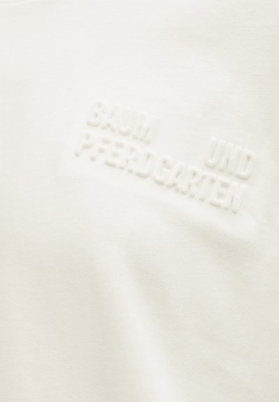 Λευκό ύφασμα με ανάγλυφο, σκαλιστό κείμενο που γράφει "BAUM UND PFERDGARTEN" με διακριτικό, τονικό στυλ.