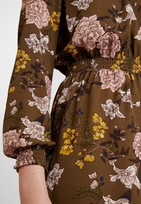Florales Kleid mit braunem Hintergrund und pinken sowie gelben Blumen. Das Design umfasst lange Ärmel mit elastischen Bündchen und eine geraffte Taille.