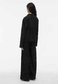 Eine Frau mit langen, dunklen Haaren trägt eine schwarze, strukturierte, florale Jacke und weit geschnittene Hosen. Sie steht mit dem Rücken zu einem schlichten weißen Hintergrund.