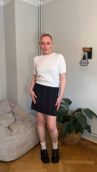 Weißes Kurzarmshirt mit gerippter Saumkante, marineblauer karierten Minirock, schwarze klobige Schuhe, weiße Knöchelsocken, mit Pflanzen und einem gemütlichen Stuhl im Hintergrund.