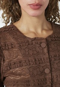 Krøllet hår person iført en brun blondetop med teksturerede flæser og to runde knapper nær udskæringen.