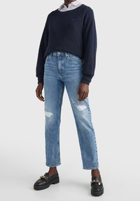 Maglione a coste blu marina indossato sopra una camicia a righe con colletto, abbinato a jeans strappati blu chiaro e mocassini neri con dettaglio in catena dorata.