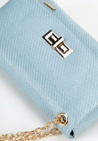 Borsa a mano intrecciata di colore azzurro chiaro con chiusura rettangolare dorata e tracolla in catena dorata su sfondo bianco.