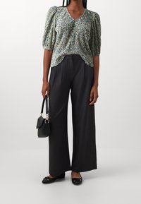 Mujer que lleva una blusa corta de manga corta con estampado floral, pantalones anchos negros, zapatos planos negros y sostiene un pequeño bolso negro.