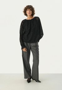 Blusa nera in pizzo con scollatura rotonda e maniche lunghe trasparenti, abbinata a pantaloni grigi chiari sartoriali. Ballerine nere a punta completano il look.