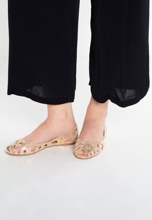 Fødder iført sorte cropped bukser og blush pink åbne flettede flade sko på et hvidt gulv.