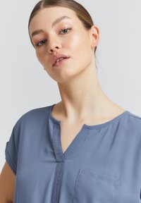 Blouse bleue à manches courtes avec un col à encolure en V, plastron avant et une poche poitrine. Tissu au toucher lisse. Coupe régulière.