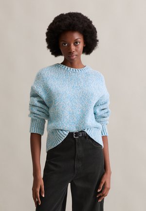 Strukturierter blauer Strickpullover mit Rundhalsausschnitt und gerippten Bündchen, kombiniert mit schwarzen, hochgeschnittenen Hosen und einem schwarzen Gürtel.