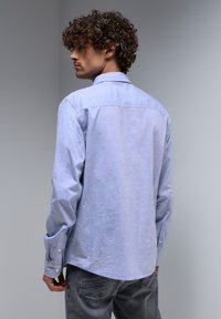 Light blue long-sleeve button-up shirt with a soft texture and curved hem. Features a standard collar and buttoned cuffs, paired with gray jeans.