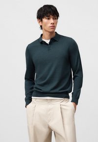 Dunkelgrüner Pullover mit Kragen und langen Ärmeln, aus einem weichen Material gefertigt. Kombiniert mit hellbeigen Hosen, die eine lockere Passform zeigen.