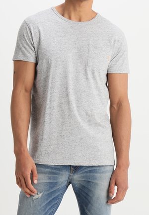 Man wearing plain light gray short-sleeve t-shirt with chest pocket and blue distressed jeans, standing against white background.