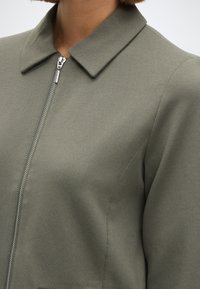 Ελαιοπράσινο μπουφάν με φερμουάρ και κολάρο, κατασκευασμένο από υφαντό ύφασμα με ορατές ραφές. Μινιμαλιστικός σχεδιασμός με κομψή εφαρμογή.